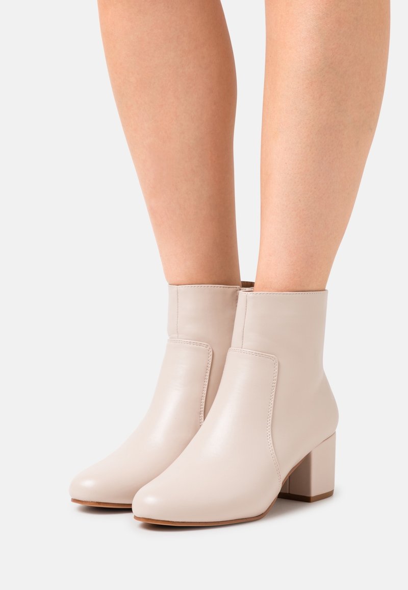 Bottes en cuir beige mi-cheville à talons carrés portées sur jambes nues sur un fond clair uni.
