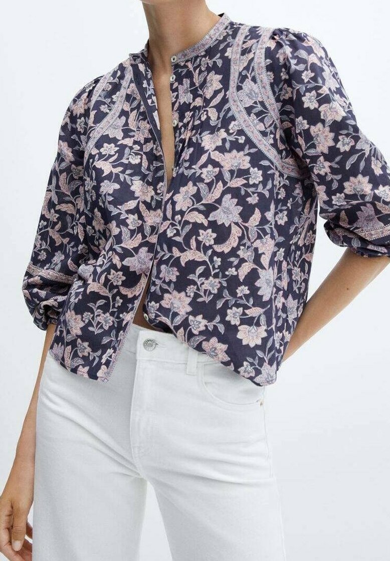 Femme portant un chemisier bleu marine à motif floral avec des manches bouffantes et un jean blanc taille haute sur un fond uni.
