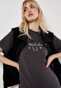 Schwarze Jeansweste über einem dunkelgrauen T-Shirt mit weißem Aufdruck, auf dem "Peace & Love CLUB Est. 2007" steht. Das Shirt hat einen runden Halsausschnitt.