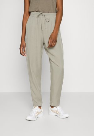 Calças jogger de cor cinza claro, de corte solto, com cintura elástica e cordão. Tecido macio com uma textura suave; combinadas com tênis brancos.