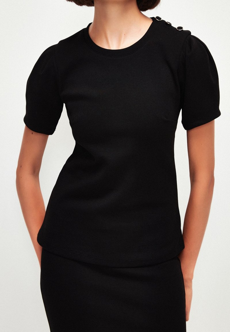 Femme portant un haut noir cintré à manches courtes avec des manches bouffantes et un bouton décoratif sur l'épaule gauche.