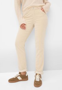 Frau trägt beige, schlanke Hosen, die an den Knöcheln aufgerollt sind, einen Cremepullover und braune Wildledersneaker mit weißen Akzenten.