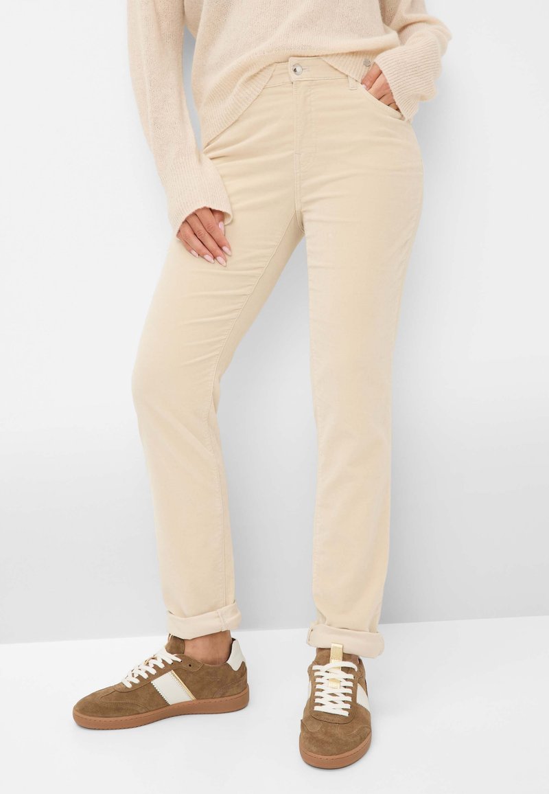 Frau trägt beige, schlanke Hosen, die an den Knöcheln aufgerollt sind, einen Cremepullover und braune Wildledersneaker mit weißen Akzenten.