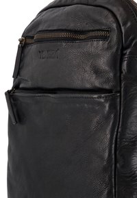 Černý kožený batoh s přední kapsou na zip, texturovaným povrchem, reliéfním logem a kontrastními kovovými zipy.