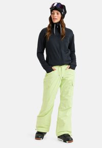 Zwarte ritsjack, cargo-stijl neon groene sneeuwbroeken en zwarte skiklaarzen. Accessoires zijn onder andere een zwarte beanie en gemirroreerde skibril.