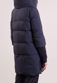 Person klädd i en lång marinblå vadderad kappa med huva och synliga ribbade ärmuppskärningar, stående mot en ljus, enfärgad bakgrund.