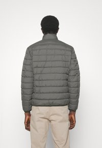 Grey puffer jacket with a quilted design, stand collar, and elastic cuffs. Worn over light beige pants, showing a smooth nylon texture.