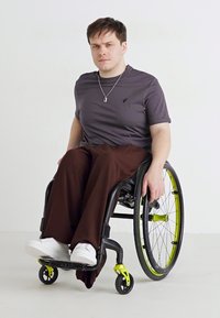 Un utilisateur de fauteuil roulant portant une chemise gris foncé et un pantalon marron. Il arbore des baskets blanches et un fauteuil roulant noir et jaune avec des roues élégantes.