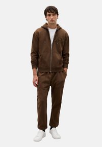 Felpa con zip marrone, dotata di tasca a marsupio e logo ricamato, abbinata a pantaloni joggers marroni e sneakers bianche. Vestibilità casual e materiale comodo.