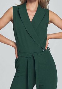 Grüner ärmelloser Jumpsuit mit Wickeldesign, der eine Bindung in der Taille und seitliche Taschen aufweist. Glatter Stoff mit einer subtilen Textur.