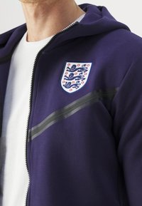 Sweat à capuche violet zippé avec une doublure blanche, arborant un emblème en forme de bouclier avec des lions, des rayures grises sur la poitrine et une texture de tissu douce.