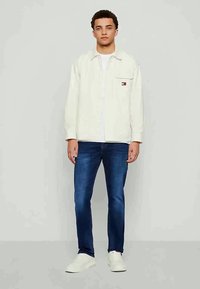 Joven de pie mirando hacia adelante, vistiendo una chaqueta blanca, jeans azules y zapatillas blancas, contra un fondo claro y liso.