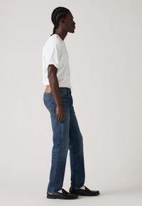 Tricou alb cu mânecă scurtă, blugi blue denim cu croială dreaptă și o pată de piele bej pe spate, și mocasini negri. Modelul stă în profil.