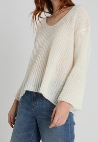 Pull léger en maille côtes fines de couleur blanc cassé avec un large décolleté, des épaules tombantes et une coupe décontractée, porté avec un jean bleu.