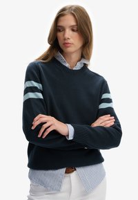 Marineblauer Strickpullover mit Rundhalsausschnitt und hellblauen Streifen an den Ärmeln. Wird über einem blau-weiß gestreiften Hemd getragen. Glatte Textur.