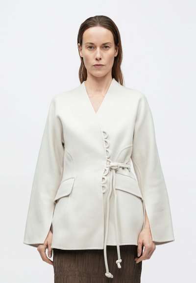 Λευκό σακάκι τύπου wrap με μακριά μανίκια, κλείσιμο μπροστά με κουμπιά toggle, πλαϊνές τσέπες και λεία υφή. Διακοσμημένο με ζώνη από παρόμοιο υλικό.