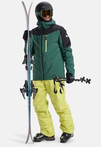 Skijacke in Grün und Schwarz mit einer Reißverschlusstasche, kombiniert mit leuchtend gelben Hosen. Skier in der einen Hand, Skistöcke in der anderen.