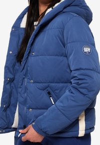 Blauwe pufferjack met een capuchon, voorzien van een fleece voering. Het heeft een rits aan de voorkant, drukknopen en contrasterende witte accenten aan de zijkanten.