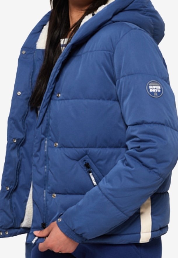 Blauwe pufferjack met een capuchon, voorzien van een fleece voering. Het heeft een rits aan de voorkant, drukknopen en contrasterende witte accenten aan de zijkanten.