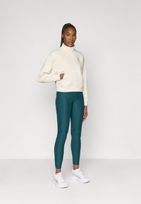 Sweatshirt de zíper em beige claro com bolso e logótipo, combinado com leggings em teal. Modelo usa sapatilhas brancas de desporto. Design simples e moderno.