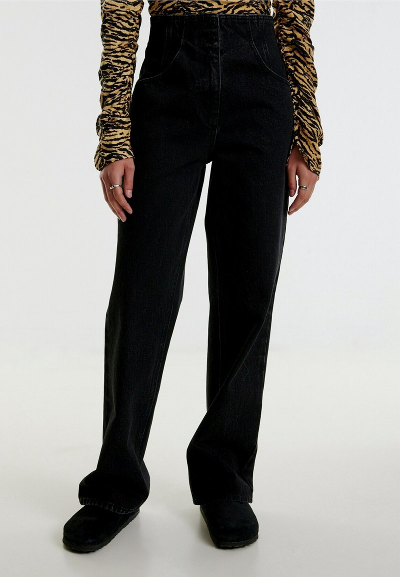 Modèle portant un pantalon noir taille haute à jambes larges, associé à des chaussures noires et un haut à manches longues imprimé tigre.
