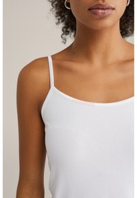 Fehér pántos top, vékony pántokkal, puha, sima anyagból készült. Mély kivágása van, és a felső test körül testhezálló.