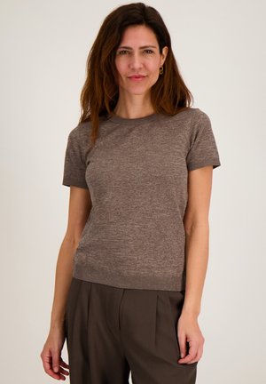 Vrouw met lang bruin haar, gekleed in een aansluitende bruine gebreide top met korte mouwen en donkerbruine geplooide broek, tegen een effen achtergrond.