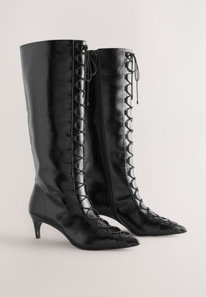 Stivali neri in pelle fino al ginocchio con punta appuntita, caratterizzati da una chiusura con lacci sul lato, tacco basso a kitten heel e chiusura con cerniera.