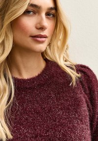 Bordowy sweter dzianinowy z teksturowanej tkaniny, z wysokim dekoltem i subtelnym połyskiem. Projekt ma miękkie, lekko bufiaste rękawy.