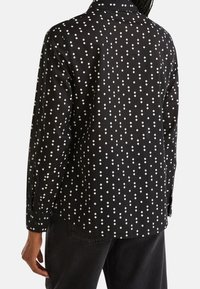 Pitkähihainen paita mustasta kankaasta, jossa on valkoinen timanttimuotoilu. Paidassa on klassinen kaulus ja nappidetaljit hihoissa.