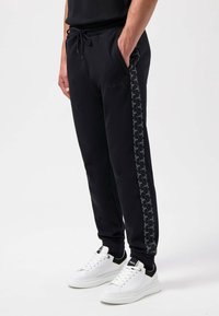 Zwarte sweatpants met een gestructureerd kettingpatroon aan de zijkant, elastische tailleband met koord en een klein zwart logo op de voorkant.