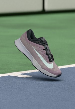 Lila-fekete Nike futócipő fehér logóval, amely a kék-zöld teniszpálya felülete fölött lebeg.