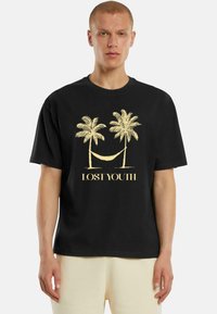 Čierne tričko s grafickým zlatým motifom dvoch paliem a hojdacej siete s textom "ZTRATENÁ MLÁDEŽ" nižšie, v uvoľnenom strihu.