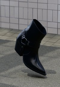 Svart ankelstövel i läder med spetsig tå, ankelrem med silverdetaljer och en klumpig klack; slät yta med polerad finish.