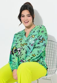 Grünbluse mit verspielten Mustern, die Totenköpfe und Pfeile zeigt, und langen Ärmeln. Kombiniert mit leuchtend gelben Hosen.