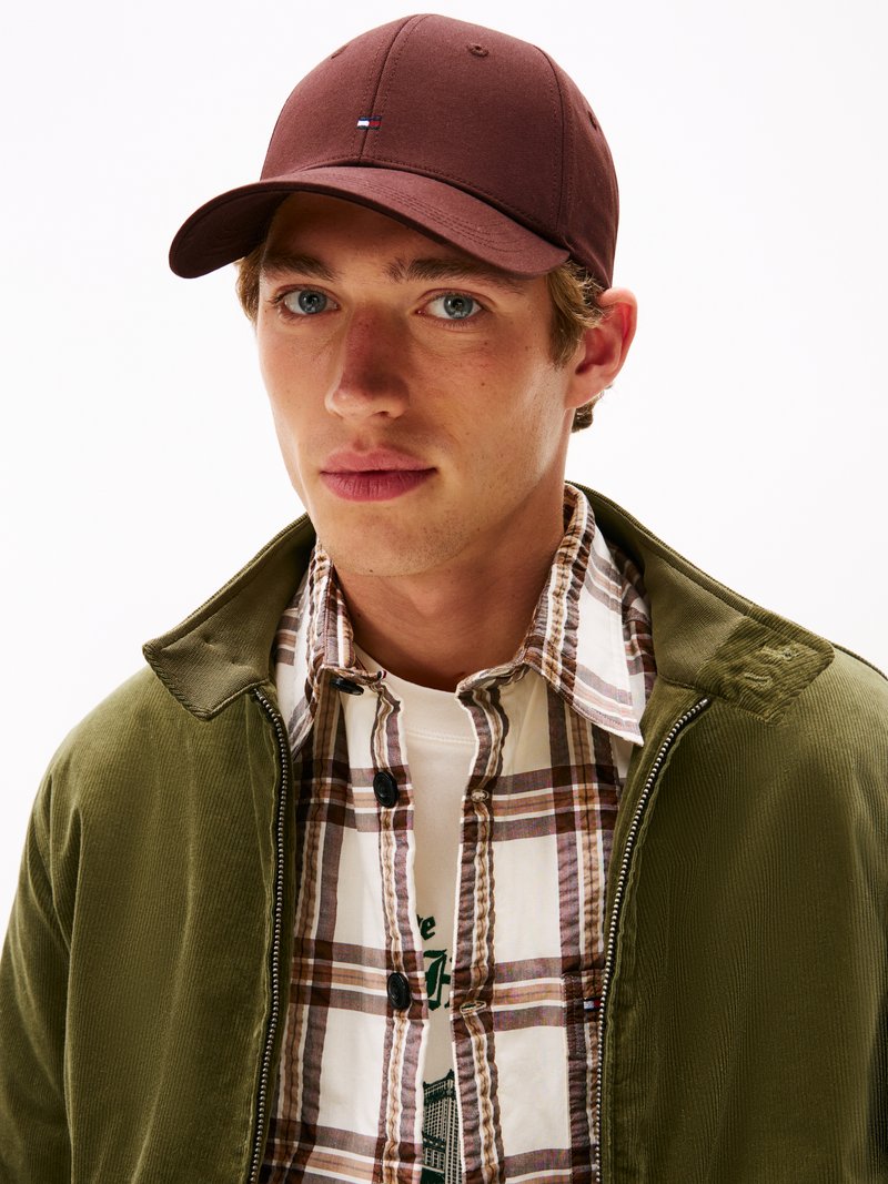Cappello da baseball marrone con logo, giacca in corda verde e camicia a quadri abbottonata sopra una t-shirt bianca. Primo piano sul volto del modello.