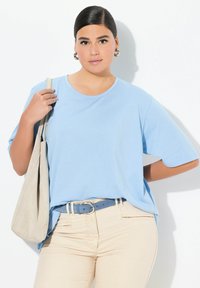 T-shirt azzurro chiaro a maniche corte, colletto rotondo, abbinato a pantaloni beige e una cintura blu. Porta una borsa a spalla beige.