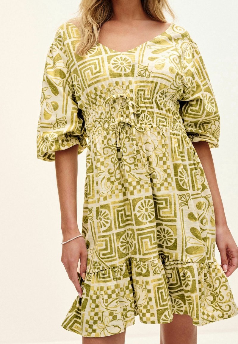 Robe à motifs verts avec manches bouffantes, taille cintrée et ourlet volanté. Présente des motifs floraux et géométriques en verts clairs et foncés.