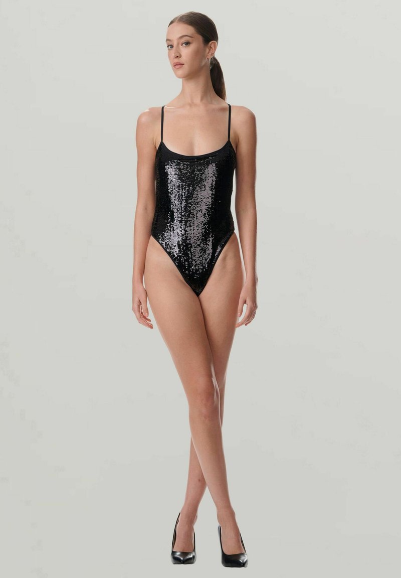 Ung kvinna står, klädd i en svart glittrande enbitar baddräkt och svarta klackskor, framför en enkel ljusgrå bakgrund.
