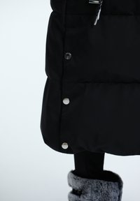 Manteau rembourré noir avec des boutons-pression argentés, présenté avec une texture lisse et un quilting visible. Le bord inférieur est légèrement arrondi, associé à des bottes grises doublées de fourrure.