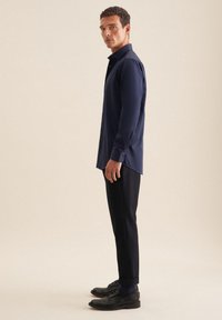 Camicia navy in misto cotone con colletto button-down, maniche lunghe e leggera lucentezza, abbinata a pantaloni scuri e scarpe nere.