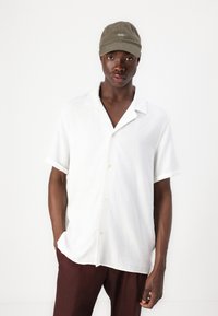 White short-sleeve shirt with a shirt collar and light texture, paired with brown lightweight trousers and a green cap featuring white text.