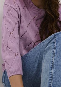 Lila gebreide trui met ruitpatronen en open steken; geribbelde halslijn en manchetten. Gecombineerd met blauwe denim jeans.