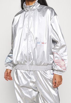 Persoon die een glanzend zilveren trainingspak draagt met een jas met ritssluiting en bijpassende broek, voorzien van rood en blauw omrande logo's op de mouwen en borst.