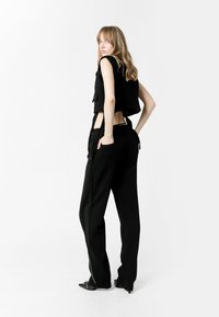 Top negro sin mangas y recortado con acentos frontales, combinado con pantalones anchos negros con bolsillos laterales y detalles con cremallera, llevados con zapatos de punta.