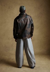 Barna és fekete márványozott bőrkabát, laza szabással, széles szürke nadrággal és zöld sapkával párosítva. Semleges háttér előtt.