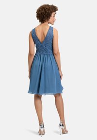 Een blauwe jurk met een kanten lijfje, V-vormige rughalslijn en een vloeiende rok. De stof is lichtgewicht, aangevuld met zilveren hakken.