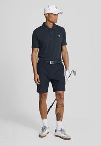 Marinefarbener Poloshirt mit kurzen Ärmeln, gelbem Logo, kombiniert mit passenden Shorts. Eine weiße Mütze, Handschuhe und ein Golfschläger runden den Look ab.