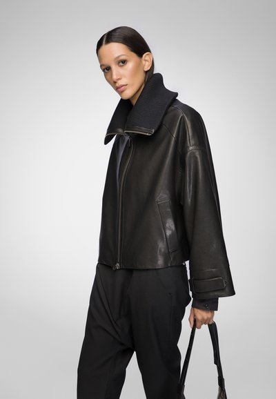 Black leather jacket with a wide, ribbed collar, front zipper, and drop shoulders, paired with loose black trousers and a handbag.