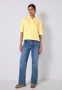 Camicia gialla di lino a maniche corte con bottoni davanti, abbinata a jeans a gamba larga blu chiaro con fessure laterali e sandali beige.